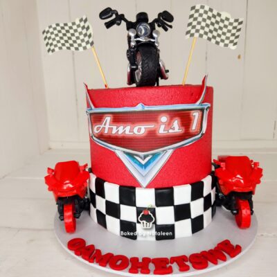 Racing-themed birthday cake with a motorcycle topper, black-and-white checkered ribbon, two checkered flags, and red decorations on a white base plate.