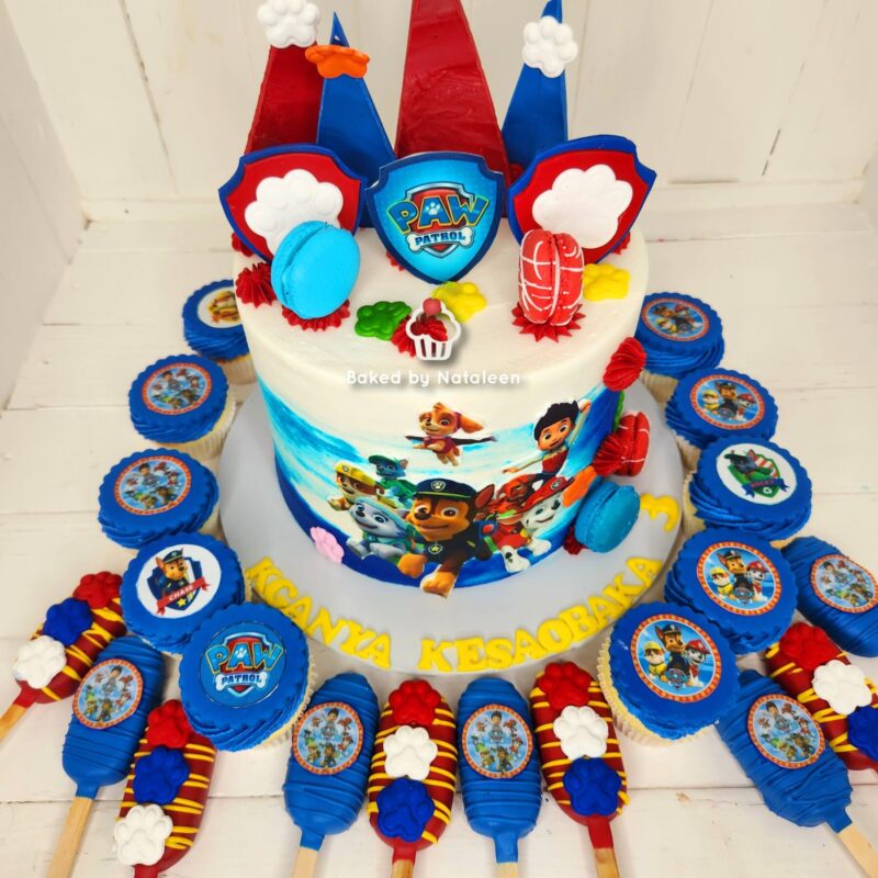 PAW Patrol themed birthday cake with blue and red toppers, character images on the side, and blue cupcakes arranged around the cake plate.
