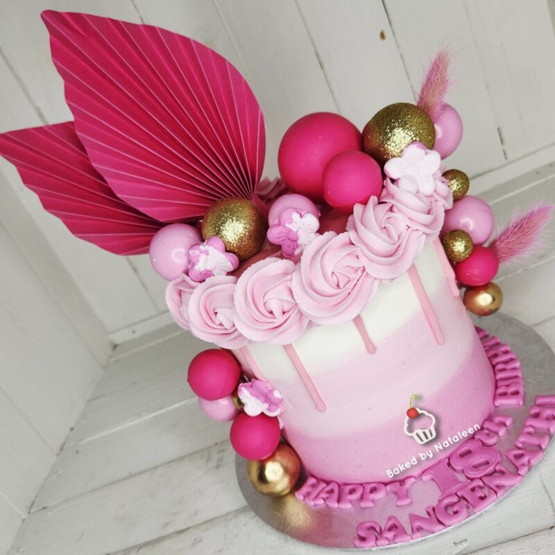 Pink and white ombre cake topped with pink rosettes, fondant balls, and gold spheres, accented by a large pink leaf.