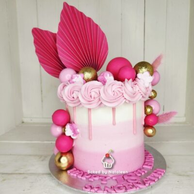 Two-tone pink and white drippy cake topped with pink rosettes, a large pink leaf, and gold and pink ornaments.