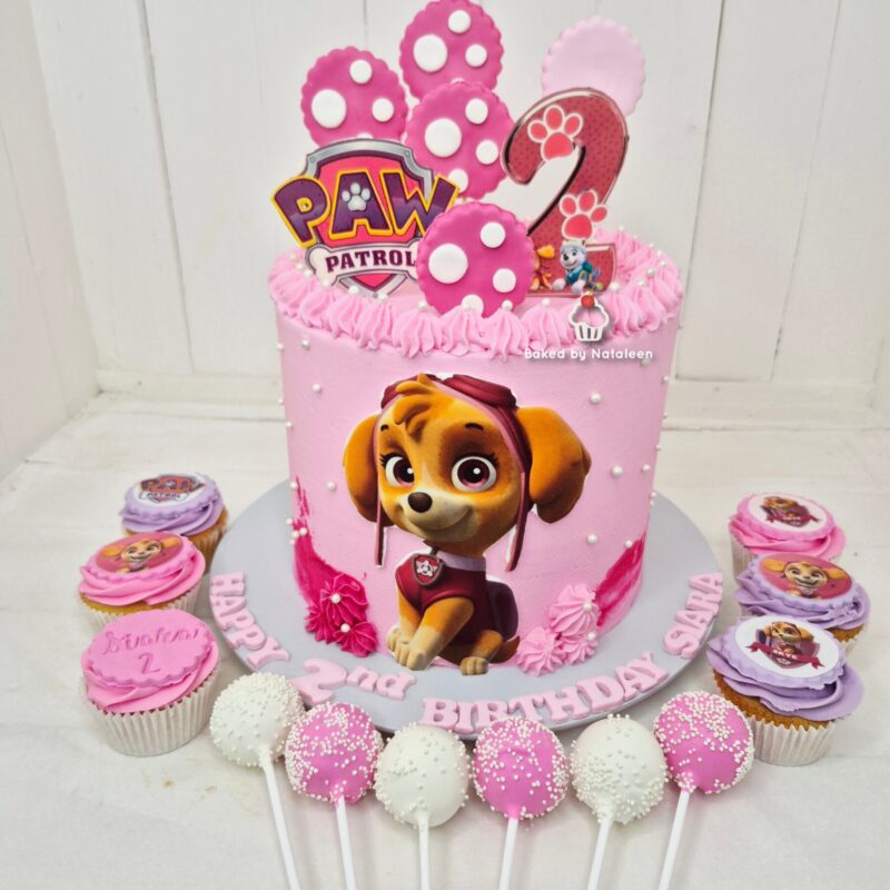 Pink Paw Patrol birthday cake with a cartoon puppy and a large number 2 topper, surrounded by cupcakes in pink and purple.