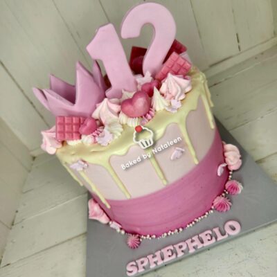 Two-tone pink birthday cake with yellow drip, large pink 1 and 2 toppers, and crown decorations for a 12th birthday. Watermark reads 'Baked by Natalieen' nearby.