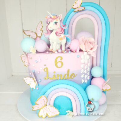 Pastel unicorn-themed birthday cake with a unicorn topper, pastel rainbow arches, butterflies, and gold '6 Lindo' on pink frosting.