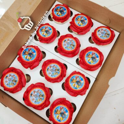 Cupcakes with red frosting and round Paw Patrol toppers arranged in a white insert box in a cardboard bakery box.