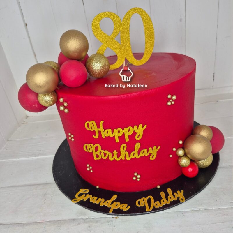 Red birthday cake decorated with gold lettering saying 'Happy Birthday', red and gold balls, and a glittery '80' topper on a black base reading 'Grandpa and Daddy' at the front.