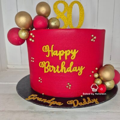 Red fondant birthday cake with gold lettering saying 'Happy Birthday' and an '80' glitter topper, adorned with gold and red spheres, on a dark board that reads 'Grandpa Daddy'.