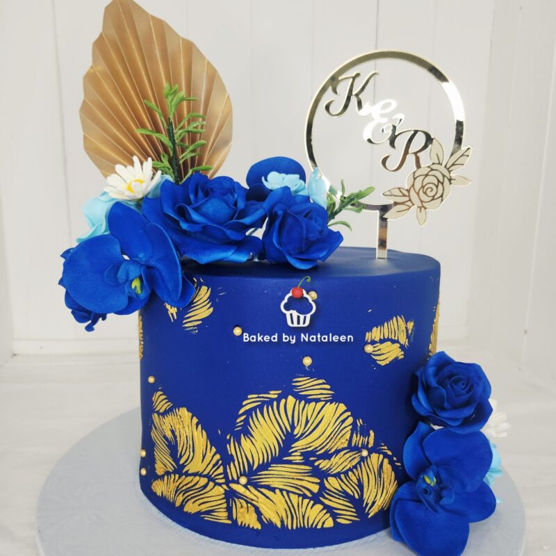 Blue fondant cake with gold leaf floral pattern, blue roses, and a circular K&R topper.