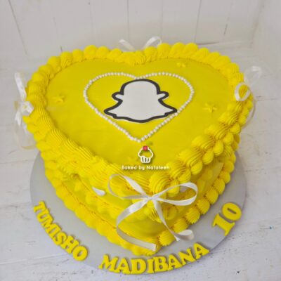 Heart-shaped yellow birthday cake with yellow piped frosting, white ribbon bows, and a Snapchat ghost emblem outlined in black on top; board reads a name and number.