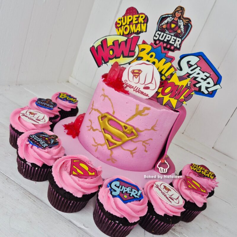 Pink superhero cake with a gold Superman emblem, surrounded by pink cupcakes with comic-style toppers on a white surface.
