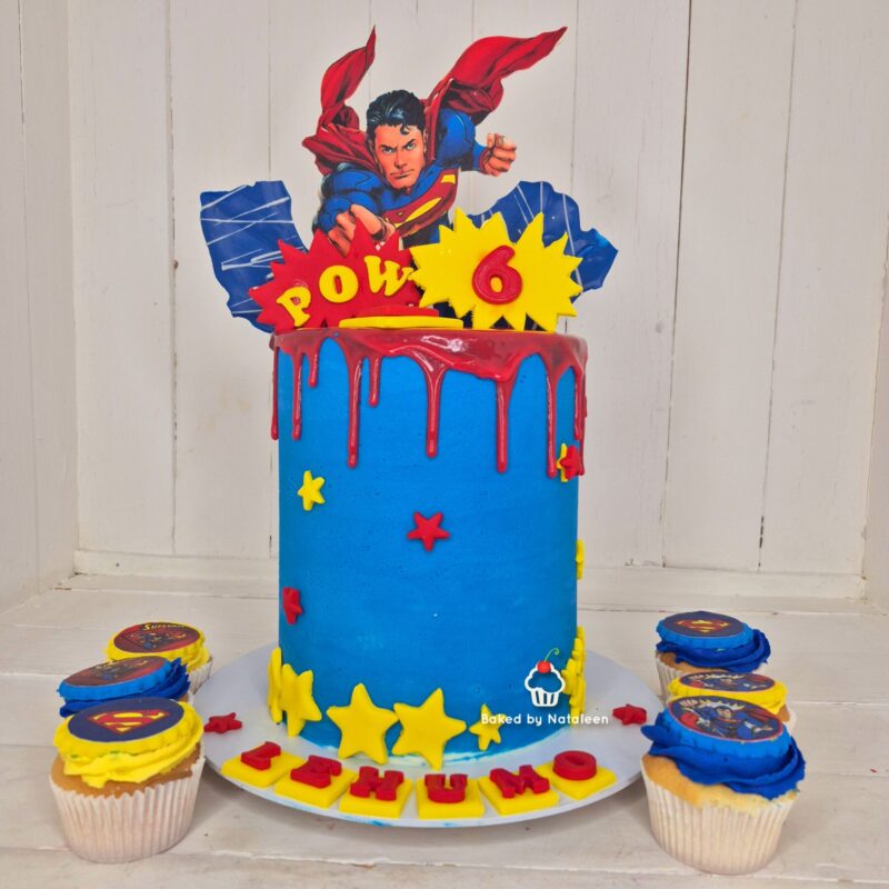 Superhero-themed birthday cake with a Superman cutout on top, blue fondant, red drip icing, and yellow stars; cupcakes surround the cake.
