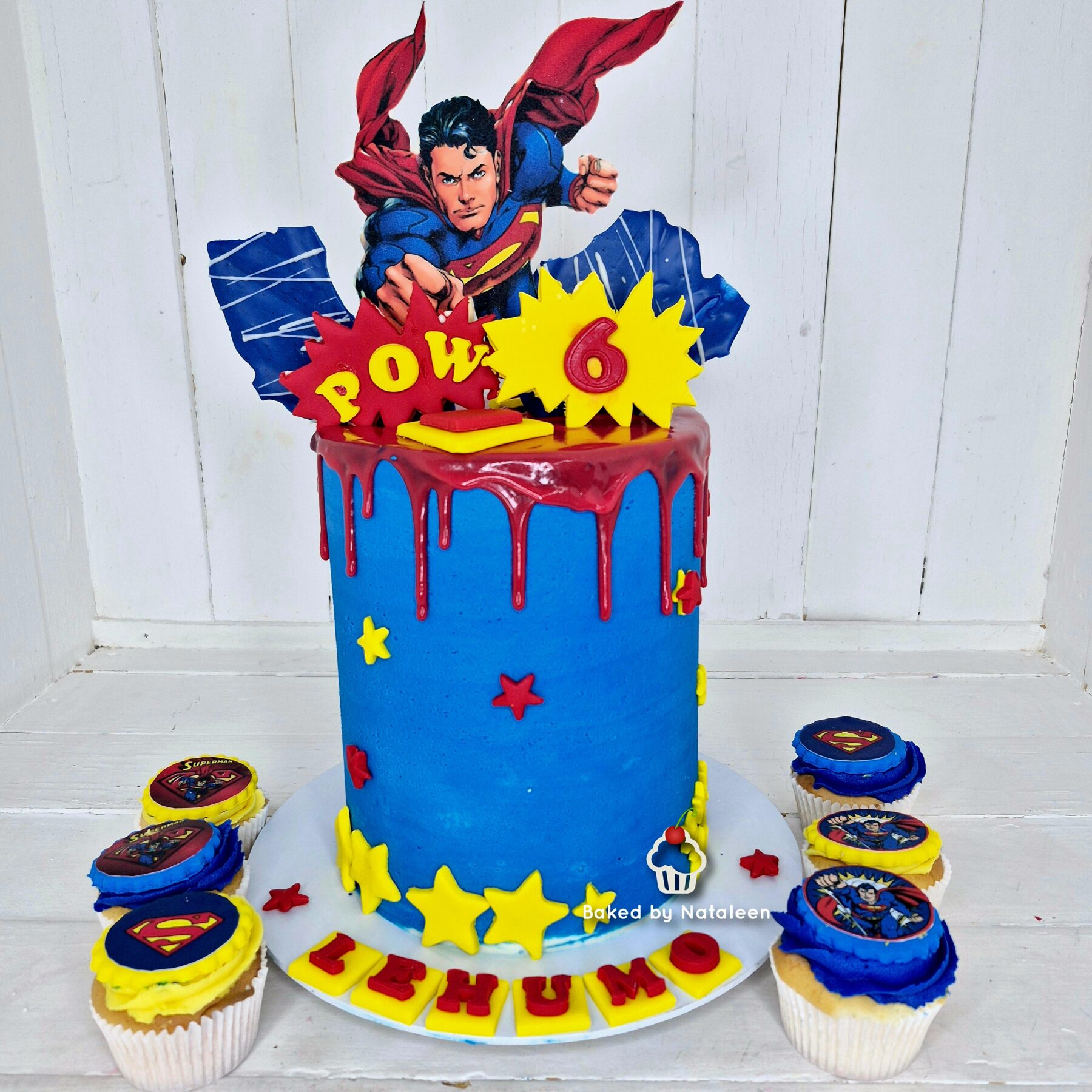 Batman? No—Superman-themed birthday cake: blue fondant, red drip glaze, yellow star decorations, and a Superman topper; cupcakes with Superman logos surround it.