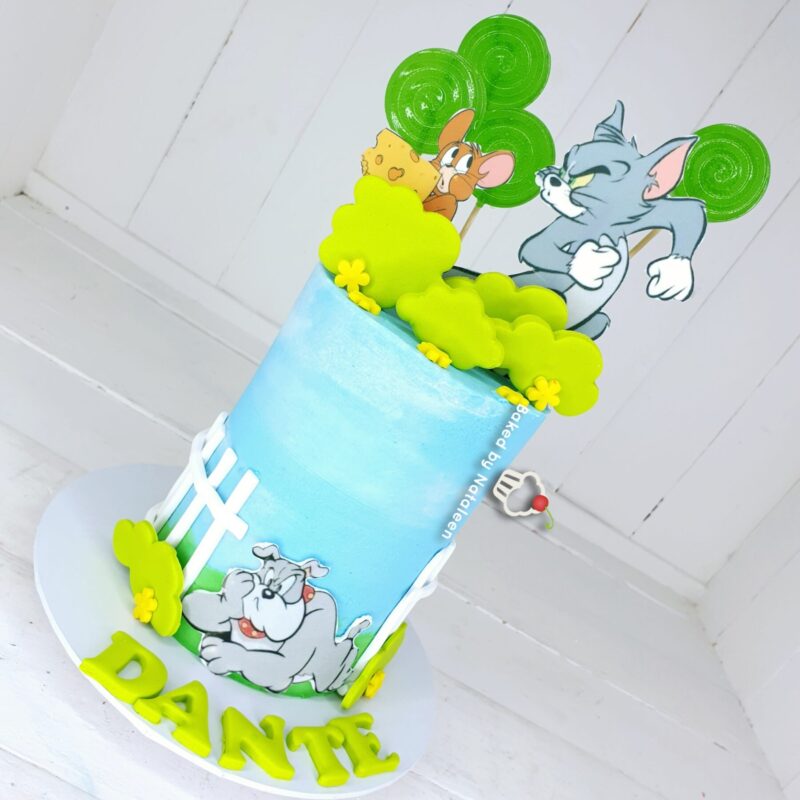 Blue birthday cake with Tom and Jerry cartoon toppers, green lollipop trees, and yellow clouds, spelling out DANTE on the base board.