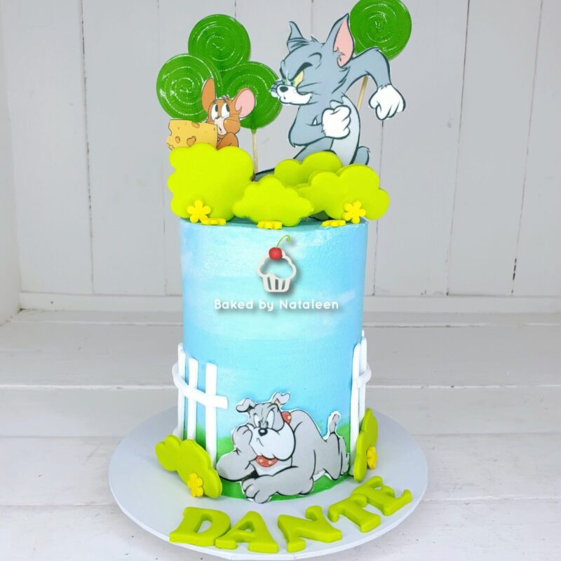 Cartoon Tom and Jerry toppers on a blue birthday cake with green trees, yellow clouds, and a white picket fence design; cake board shows large yellow letters spelling a name.