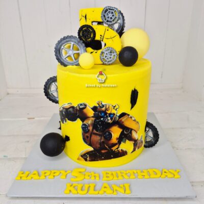 Yellow Transformer-themed birthday cake with gear toppers and the message 'Happy 5th Birthday Kulani' on the base board.