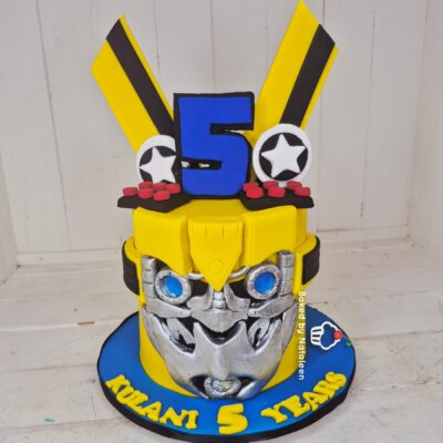 Birthday cake sculpted as a yellow armored figure with a blue number 5 on the chest, silver gears, and a blue base plate.