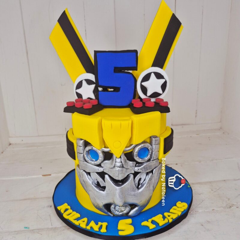 Birthday cake sculpted as a yellow armored figure with a blue number 5 on the chest, silver gears, and a blue base plate.