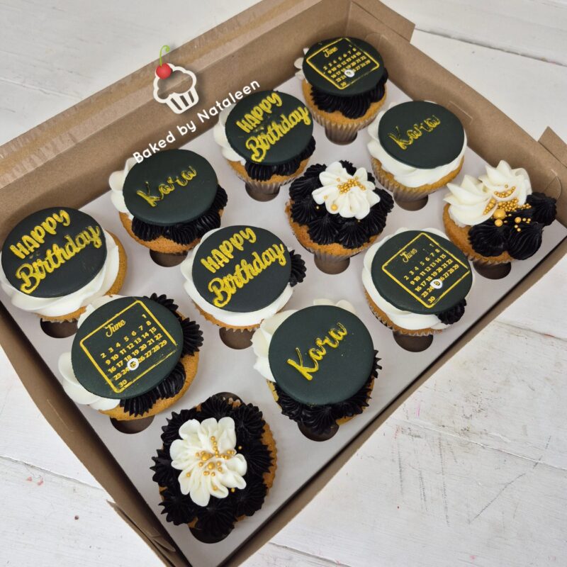 Box of twelve cupcakes in a cardboard bakery box, white frosting with black and gold toppers reading 'Happy Birthday' and decorative flowers.