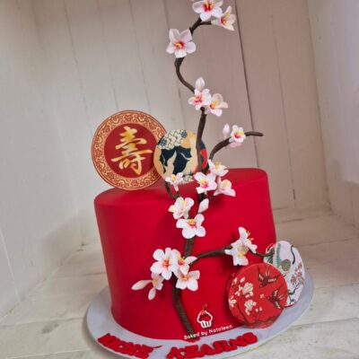 Red cake adorned with a plum blossom branch and decorative round ornaments, including a gold coin with the Chinese 'shou' character, on a white base.