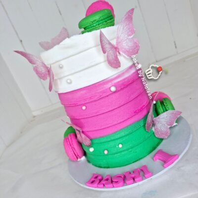 Three-tier cake with white, pink, and green fondant, pink macarons, pearl decorations, and pink butterfly accents on a white base reading BASHY.