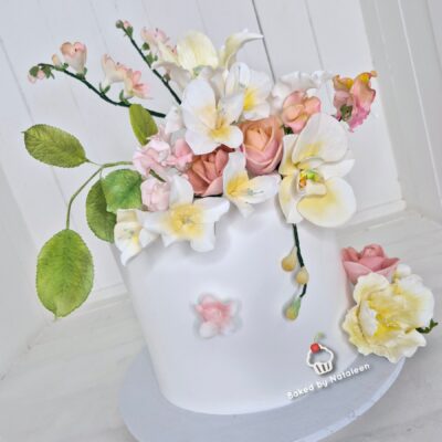 White fondant cake topped with pastel sugar flowers and roses, with green leaves and a light background.