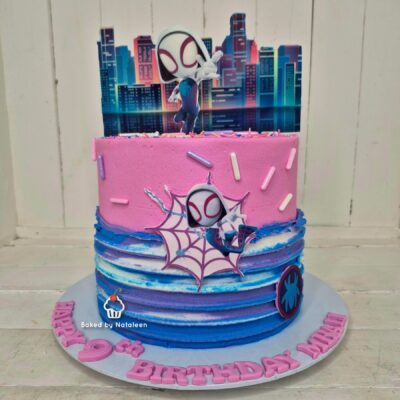 Birthday cake with pink and blue layers, topped by a Spider-Man figure hero against a cityscape backdrop; pink sprinkles on top and decorative spider-themed cutouts on the sides; partial 'Birthday' lettering around the cake board.