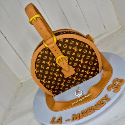 Round handbag-shaped cake with a brown Louis Vuitton monogram pattern and tan straps on a light blue platter