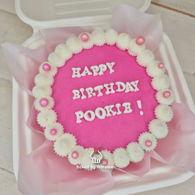 Round pink birthday cake with white piped frosting border and pink sugar pearls, placed in a white box with tissue lining; icing reads, 'HAPPY BIRTHDAY POOKI!'.