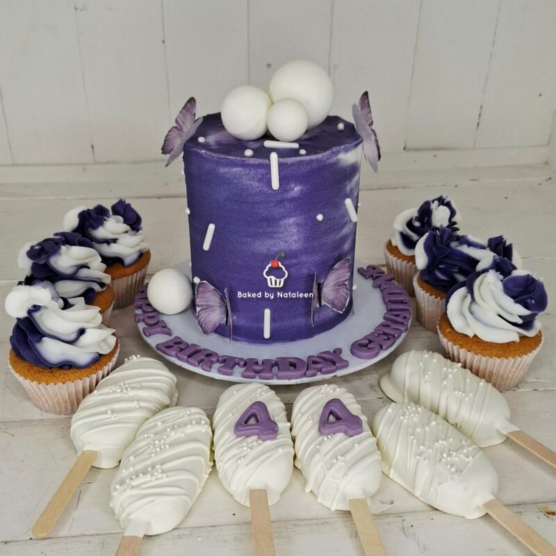 Purple birthday cake with butterfly decorations and white fondant balls, surrounded by matching cupcakes and white chocolate pops with a purple '4' motif on a white tablecloth.