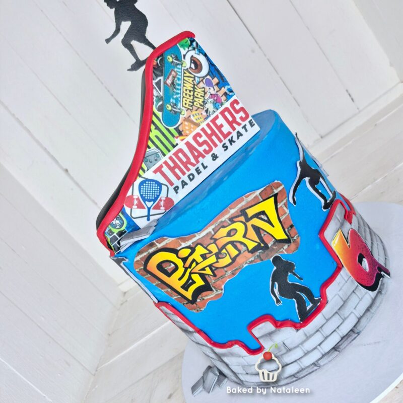 Two-tier birthday cake covered in blue and gray with skate‑and‑gaming logos (Thrasher, Diggin’, etc.), brick street detailing, and a small cupcake topper at the base.