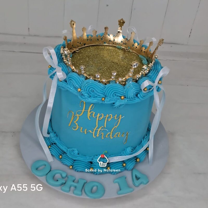 Blue birthday cake with a gold crown topper, turquoise icing, white ribbons, and gold accents; 'Happy Birthday' written on the side.