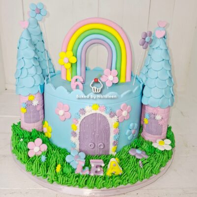 Whimsical birthday cake with two blue castle towers, a central pastel rainbow, flowers, and the name LEA on a green grass base.