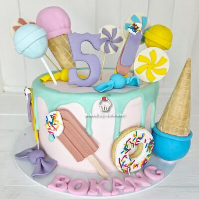 Pastel birthday cake with turquoise drip, topped by a purple ‘5’ and assorted candy toppers, including lollipops, an ice cream cone, a sprinkled donut, and a chocolate bar.