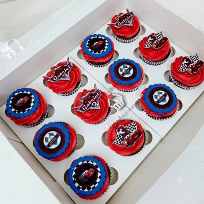 Box of twelve red-frosted cupcakes with blue accents and racing-themed toppers inside a white display box.
