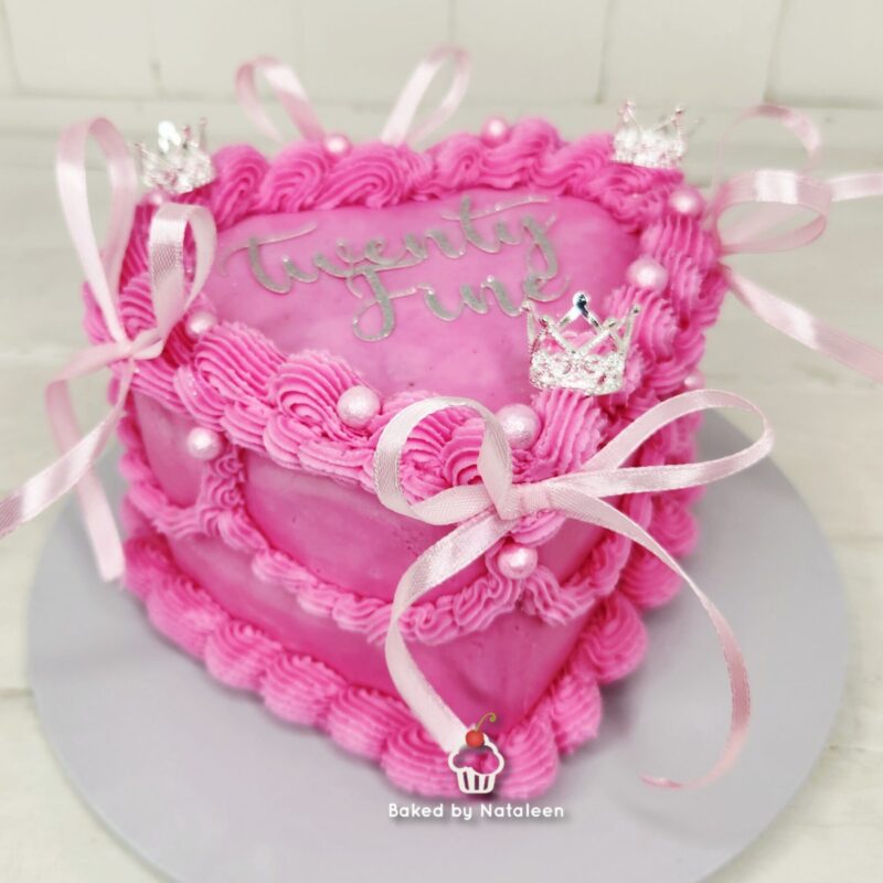 Pink, princess-themed cake with decorative swirls, ribbons, and tiny crowns; text 'Baked by Natalee n' at the base.