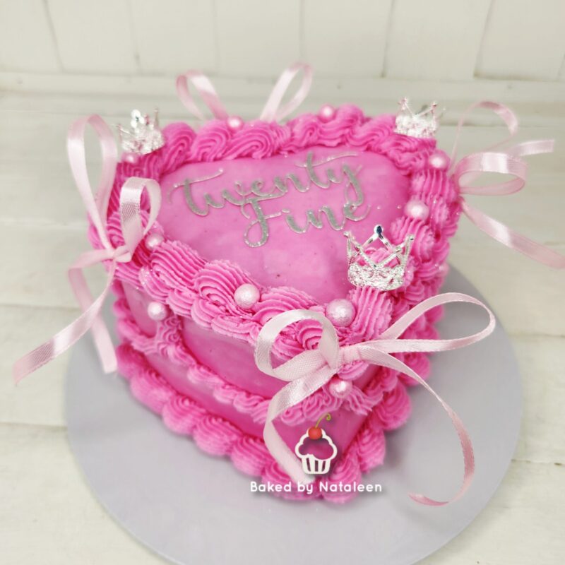 Pink heart-shaped birthday cake with rosette frosting, pink ribbons, pearls, and small crown decorations, with 'twenty five' written in silver.