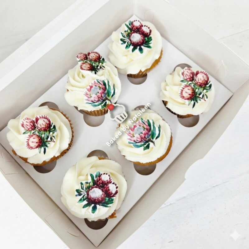 Six cupcakes in a white box, each with swirled vanilla frosting and pink-red floral toppers.
