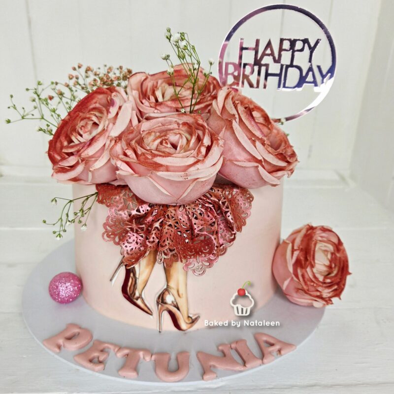 Pink birthday cake with a bouquet of large pink roses, delicate greenery, and a round 'Happy Birthday' topper on a pale pink base.