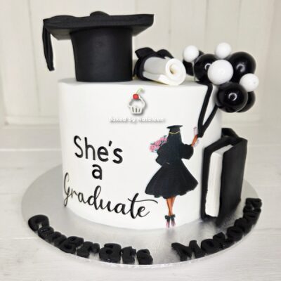 Graduation cake with 'She's a Graduate' message, black cap, diploma, and balloon decorations on a white base.