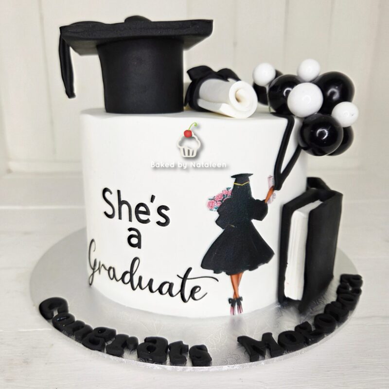 Graduation cake with 'She's a Graduate' message, black cap, diploma, and balloon decorations on a white base.