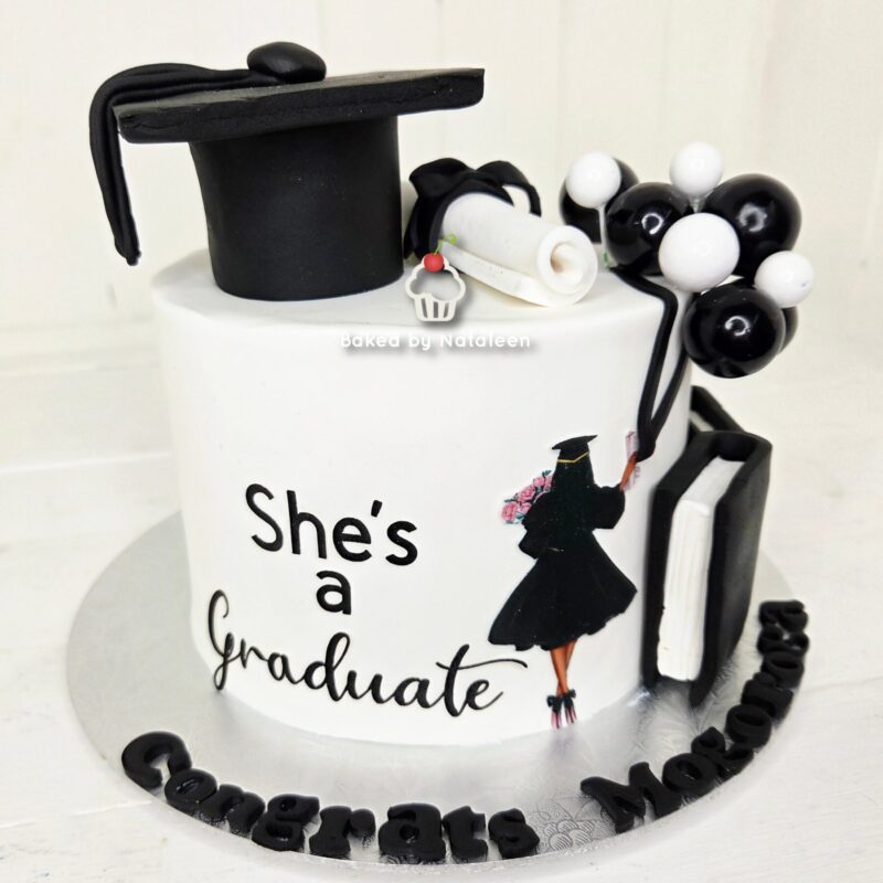 Graduation cake with a black mortarboard topper, the words "She's a Graduate" and a silhouette of a graduate on the side, plus a bouquet of black-and-white balloons and a book-shaped tower on the right.