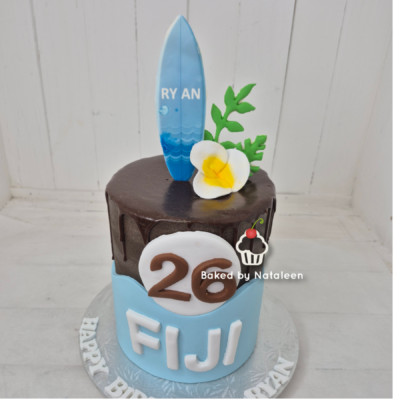 Two-tier birthday cake with chocolate top, blue bottom tier reading FIJI, a white-number 26 emblem, and a surfboard topper with 'RYAN' and a white flower.