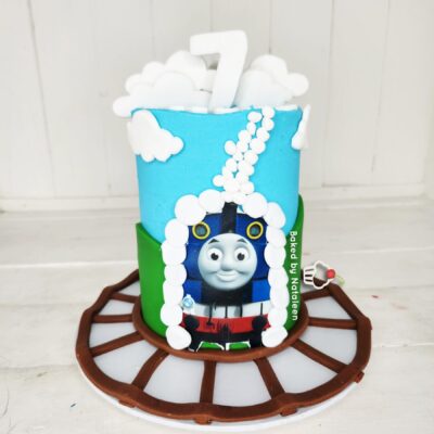 Three layer Thomas the Tank Engine birthday cake with blue and green fondant, white clouds, and a large white 7 topper on a brown track base.