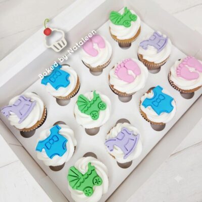 Twelve cupcakes with white frosting and pastel baby-themed fondant toppers (blue and purple onesies, pink bows, green leaves) in a white box.