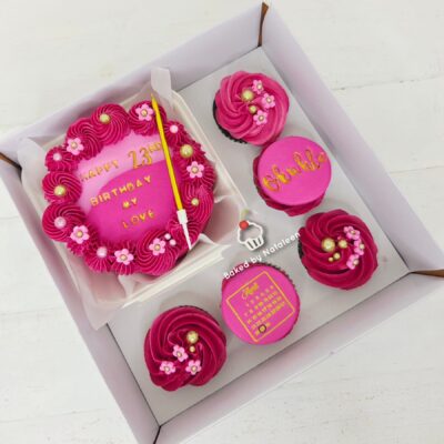 Pink birthday cake with flower decorations and a candle in a white box, surrounded by pink-frosted cupcakes on a tray.