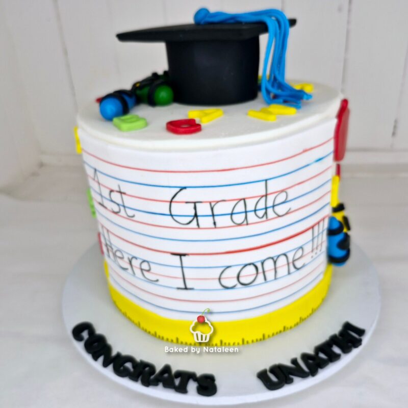 Graduation-themed cake with a black graduation cap and blue tassel on top, colorful fondant letters around the side spelling 'First Grade', and a yellow base.