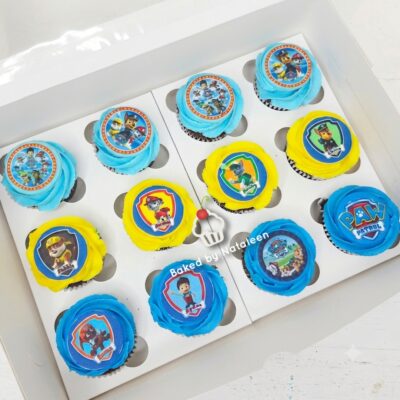 Twelve Paw Patrol cupcakes in a white box with blue and yellow frosting and character toppers, arranged in a tray.