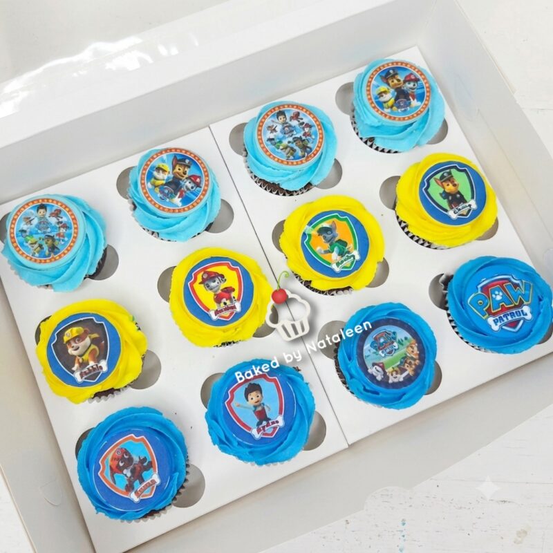 Twelve Paw Patrol cupcakes in a white box with blue and yellow frosting and character toppers, arranged in a tray.