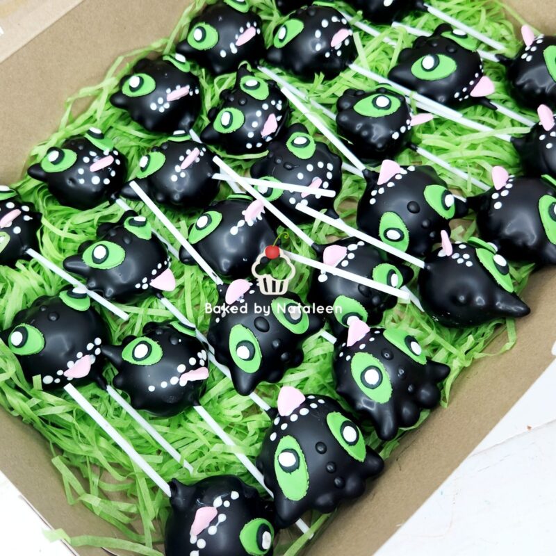 Black cat/monster-shaped cake pops with bright green eyes and pink ears, arranged on white sticks in green paper filler within a box.