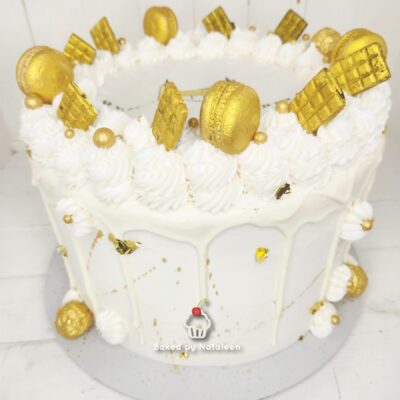 White frosted cake adorned with gold macarons, gold wafers, and piped white icing swirls along the edge.